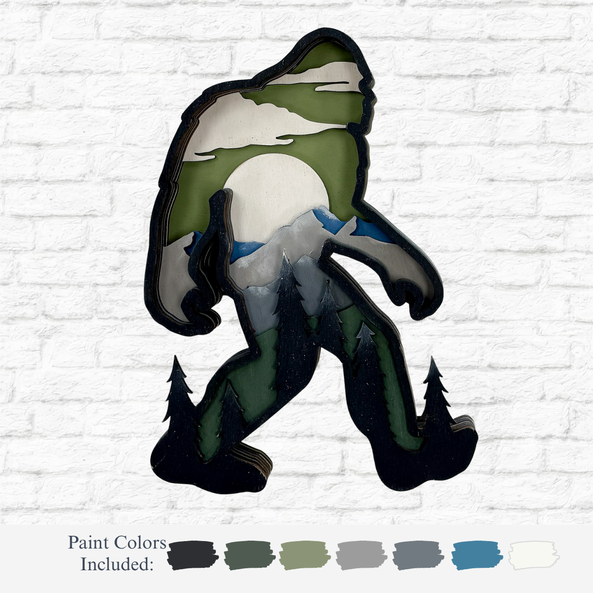 The Rustic Rhode’s DIY 3D Bigfoot Kids Kit includes a Baltic Birch Bigfoot cutout, painted with green trees, gray mountains, a white sun, and clouds, plus six paint colors on a white brick background.