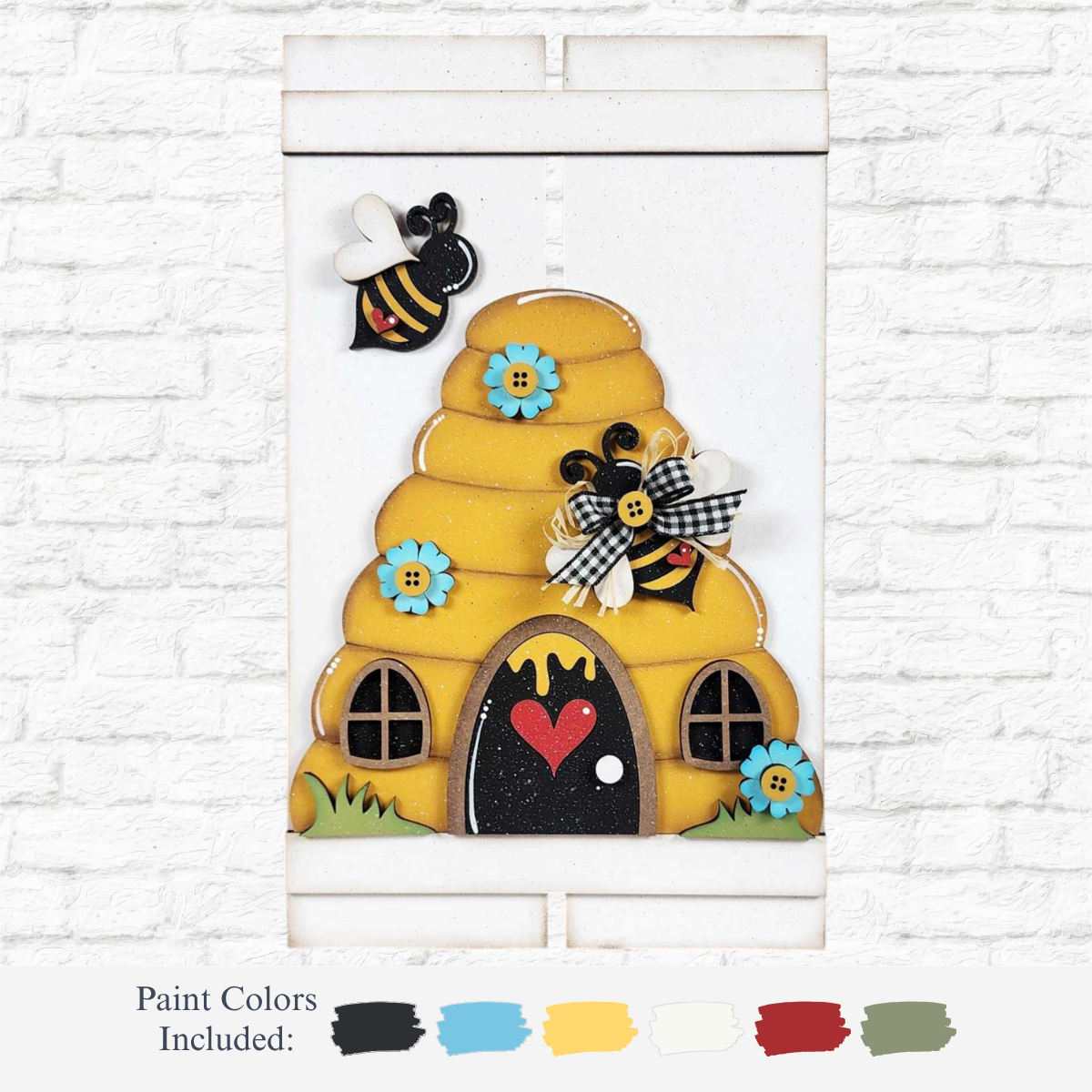 The Rustic Rhode’s DIY Beehive Pallet Sign features two bees, blue flowers, bows, and a red heart on wood against a white brick wall, with included paint color swatches—a charming handmade décor piece.