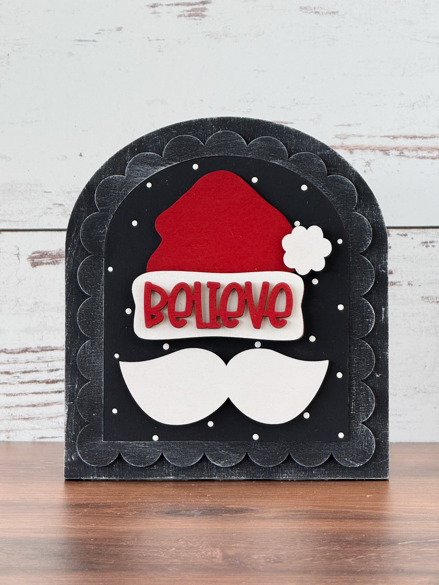 The Rustic Rhodes DIY Scalloped Arch Insert Frame features a red Santa hat and white mustache on a black scalloped background with BELIEVE in red letters and white snow-like dots, displayed on a wooden surface.