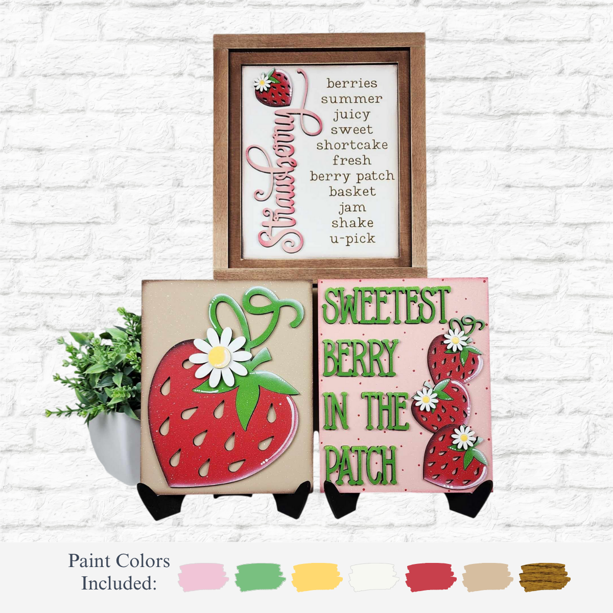 Three strawberry-themed signs from The Rustic Rhode DIY Berry Sweet Sign Trio are displayed against a white brick wall, accompanied by two paint pots, green leaves, and swatches of seven paint colors below.