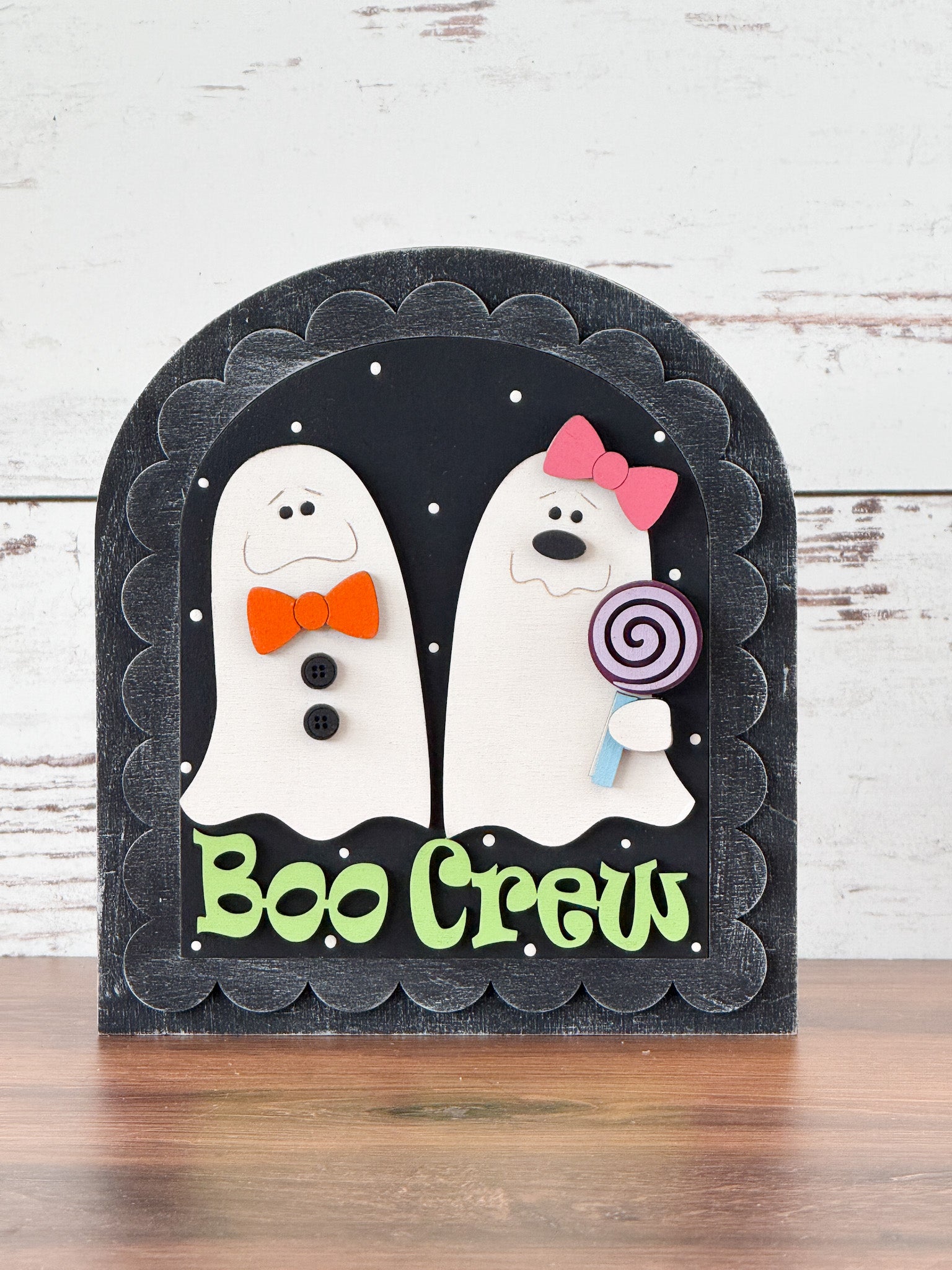 The Rustic Rhode’s DIY Scalloped Arch Insert Frame displays two cartoon ghosts with bows and buttons—one holding a lollipop. Boo Crew is written in green, and the black background with white dots resembles stars.