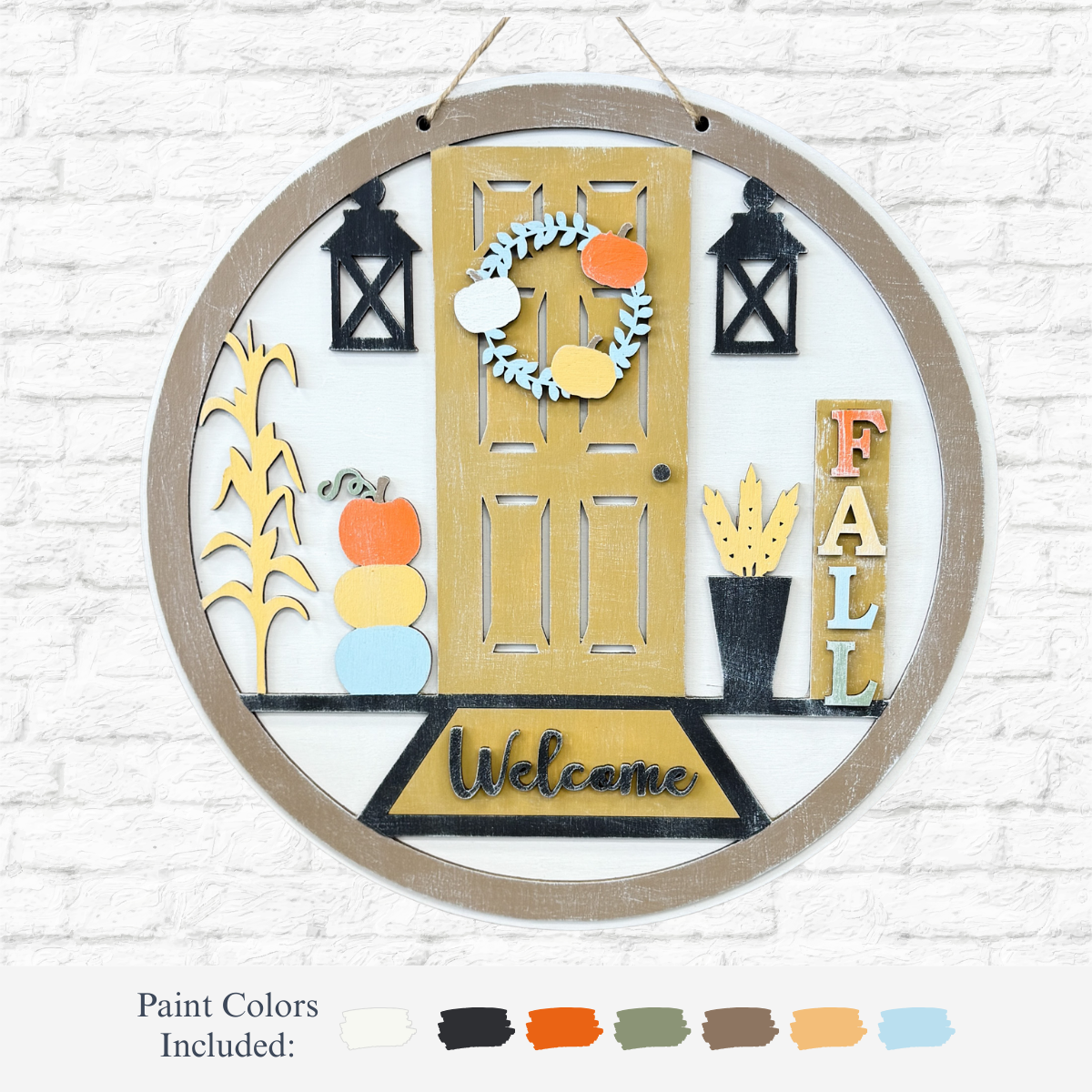 The Rustic Rhode DIY Cozy Autumn Porch Door Hanger features a round wooden sign with yellow door, fall wreath, lanterns, pumpkins, FALL sign, potted plant & welcome mat—perfect for autumn porch decor. Paint samples shown on white brick background.