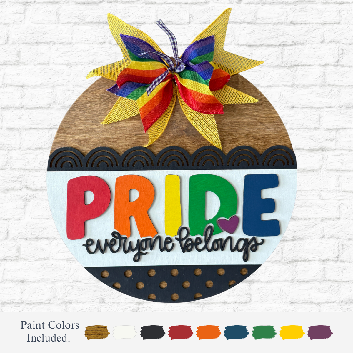 The Rustic Rhode DIY Everyone Belongs Door Hanger features a round laser-cut wood shape, a colorful bow, “PRIDE” in rainbow letters, and “everyone belongs” in script. Shown on a white brick wall with paint colors for the kit displayed below.