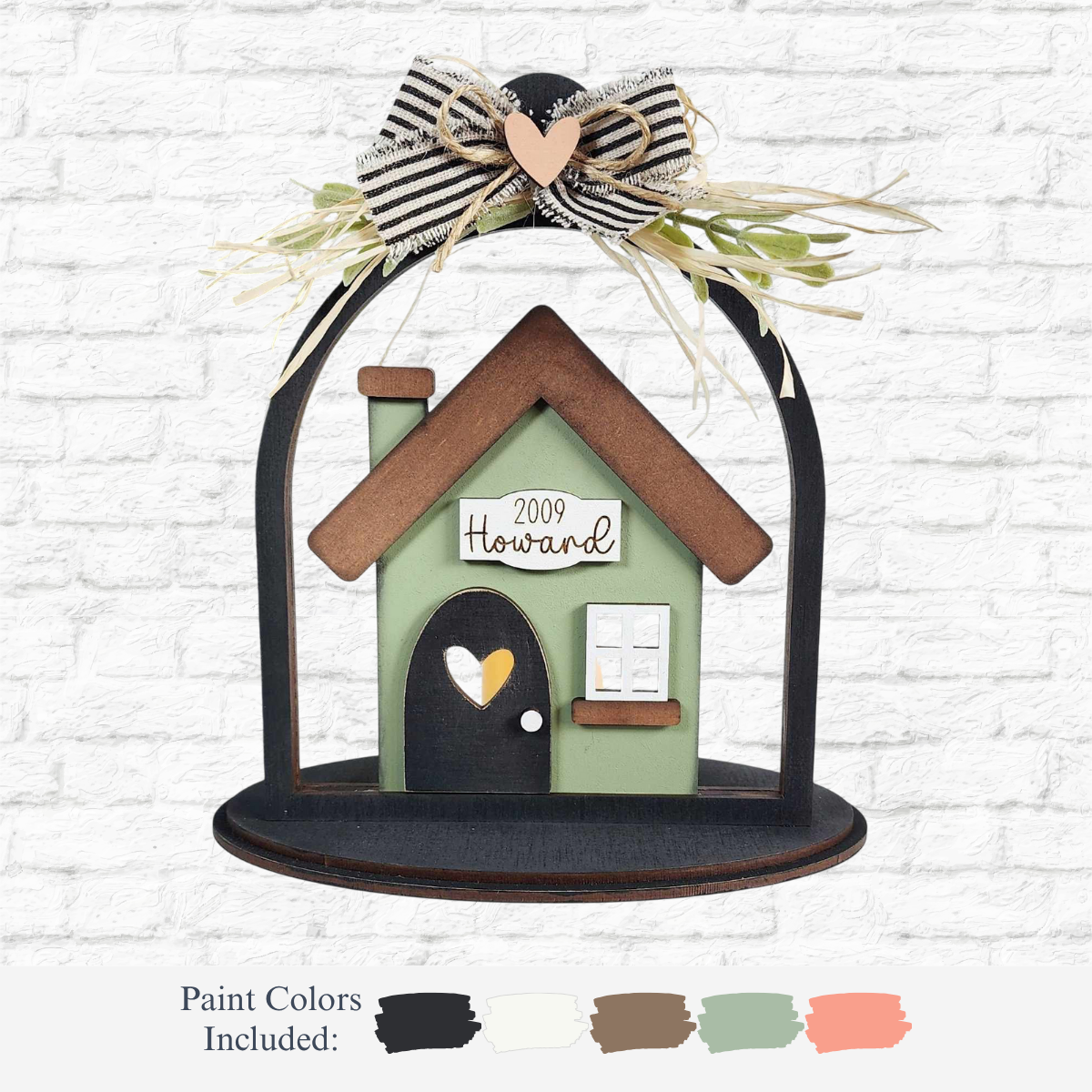 The Rustic Rhode’s DIY Family Home Cloche Votive Holder is a personalized home décor kit with a green base, brown roof, white accents on a black stand, striped bow with greenery, and paint swatches in black, white, brown, green & orange.