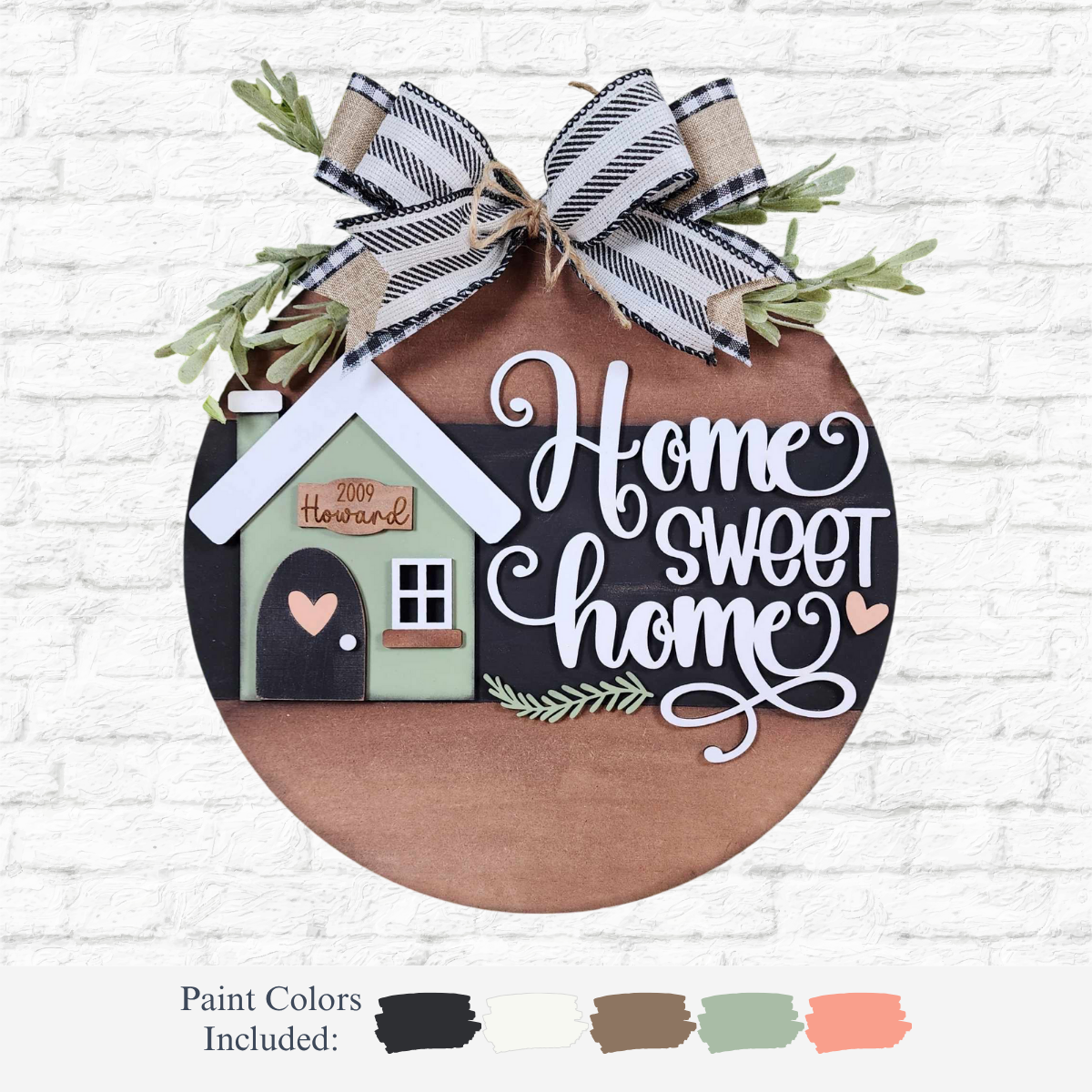 The Rustic Rhodes DIY Family Home Door Hanger is a round wooden sign with a black and white plaid bow, greenery, and house detail. Home sweet home is scripted above five paint color choices labeled with their names.