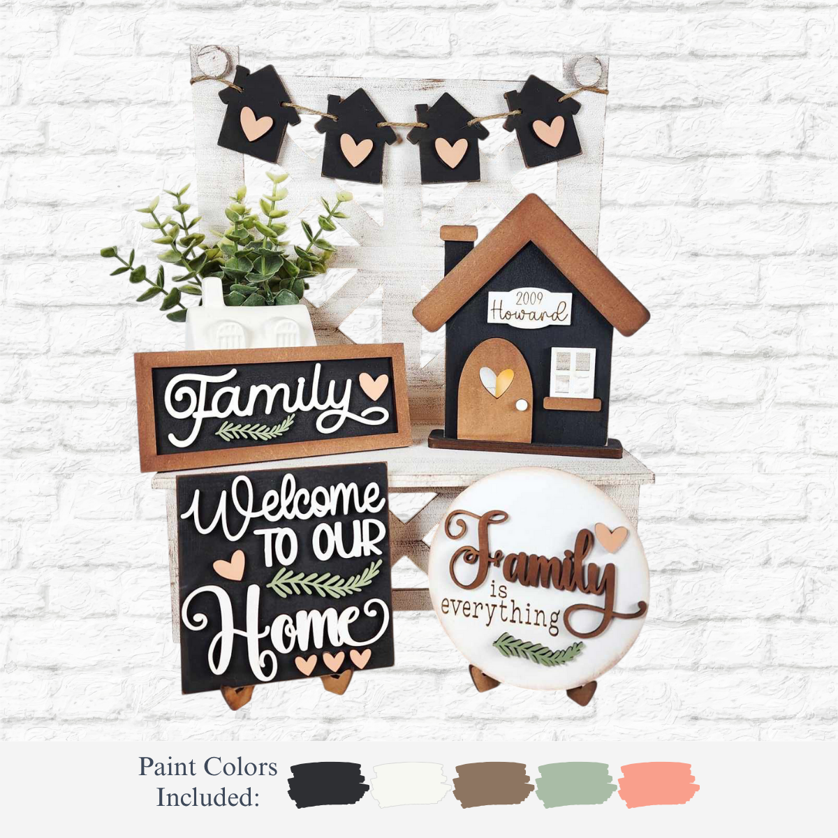 The Rustic Rhode’s DIY Family Home Tiered Tray Kit includes signs saying Family, Welcome to our Home, and Family is everything, plus a heart, leaf motifs, small house, faux plant, and mini house garland on a white brick background.