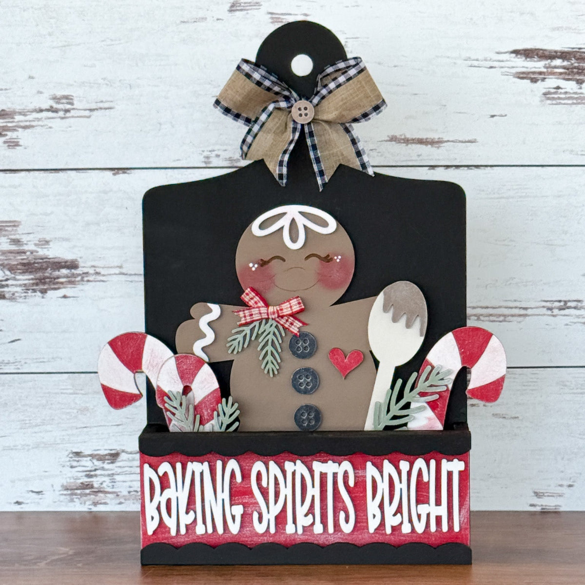 The Rustic Rhode’s DIY Interchangeable Breadboard Frame features a gingerbread figure, candy canes, greenery, and a wooden spoon in a “Baking Spirits Bright” box with a black and burlap bow, set on a white wooden background.
