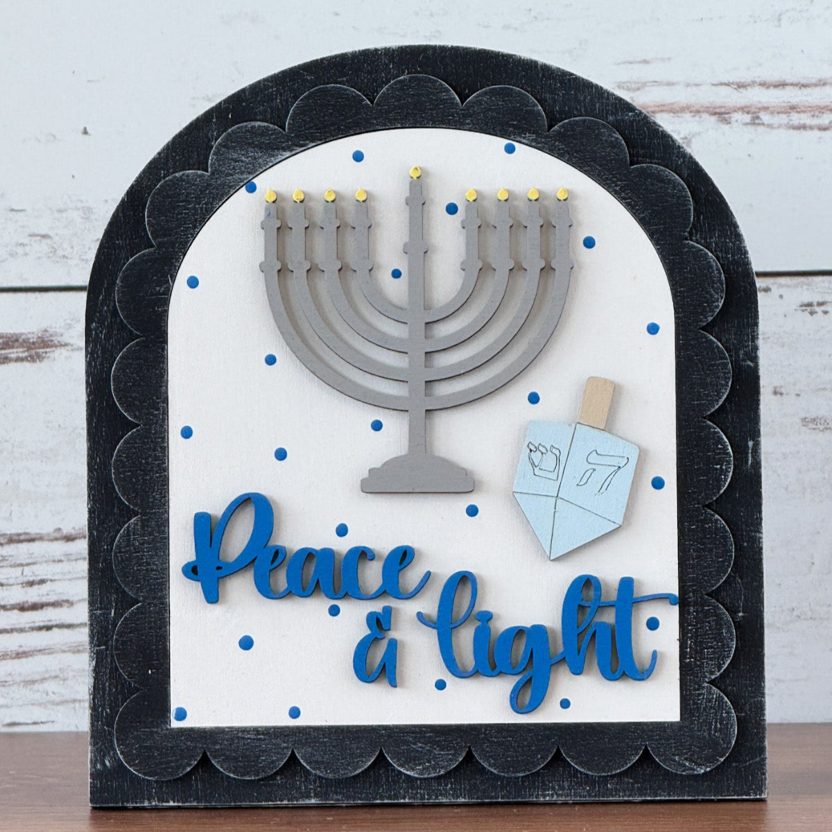 The Rustic Rhode DIY Hanukkah Peace & Light Arch Insert features a gray menorah, blue dreidel, and “Peace & light” in blue script on white with blue dots—perfect for Hanukkah crafts or unique holiday décor.