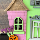 A close-up of The Rustic Rhode's DIY Haunted House Kit features a pink house with green trim, a black cat at the window, purple flowers, and an orange pumpkin in front.
