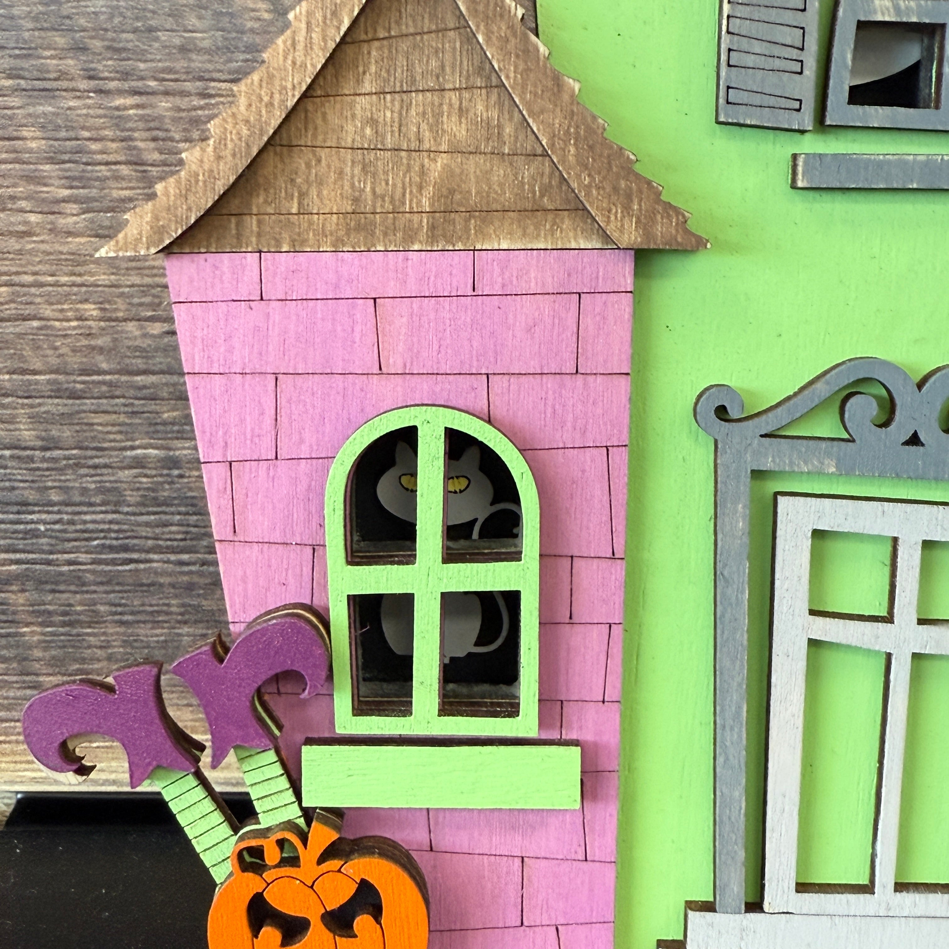 A close-up of The Rustic Rhode's DIY Haunted House Kit features a pink house with green trim, a black cat at the window, purple flowers, and an orange pumpkin in front.