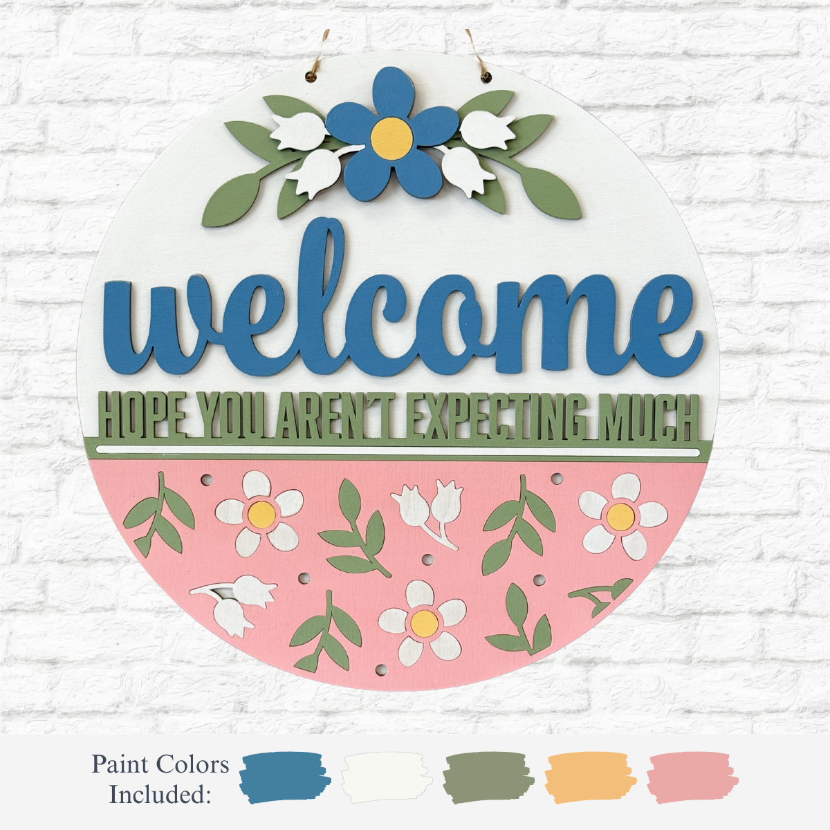 The Rustic Rhode’s DIY Hope You Weren’t Expecting Much Door Hanger features “welcome” in blue and “hope you aren’t expecting much” in green, with floral accents on a pink and white background—perfect for a creative door project.