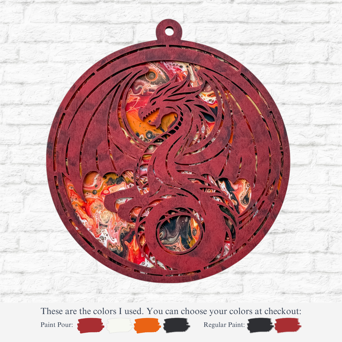 The Rustic Rhode’s DIY Paint Pour-Dragon kit features a circular ornament with a cutout dragon, painted in red, black, orange, and white using the pour technique. Paint color options are displayed below against a white brick wall background.