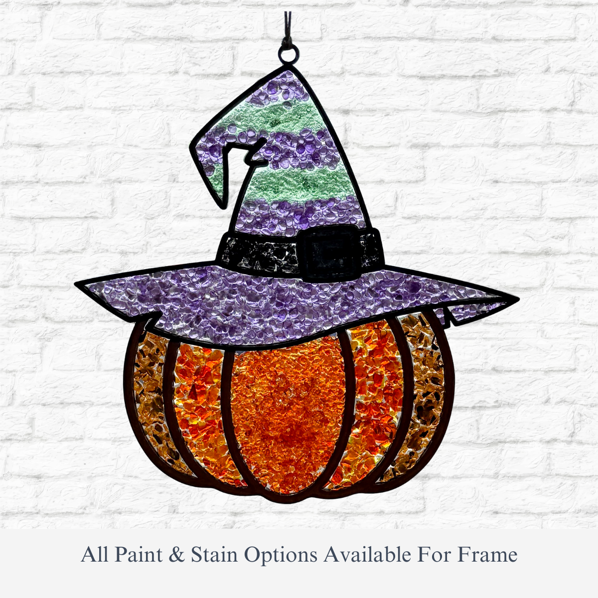 The Rustic Rhode DIY Pumpkin Witch Faux Suncatcher, shaped like a pumpkin in a striped purple witch hat, hangs on a white brick wall as festive Halloween window decor. All paint and stain options available for the frame.