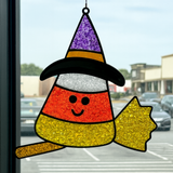 The Rustic Rhodes DIY Candy Corn Witch Faux Suncatcher hangs on a window, shaped like a smiling candy corn with a witchs hat and broom, with views of a parking lot and buildings outside.