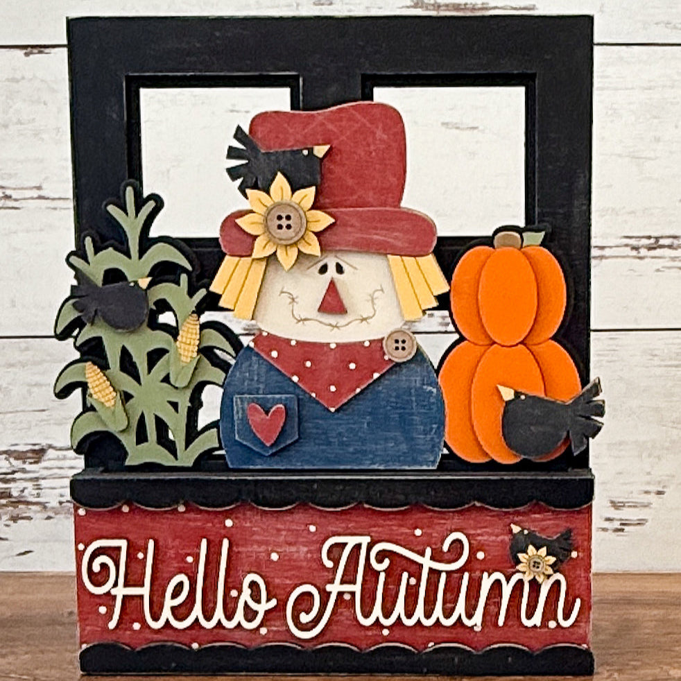 The Rustic Rhode’s DIY Interchangeable Window Frame features a decorative sign with a cheerful scarecrow, sunflowers, corn, pumpkins, blackbirds, and “Hello Autumn” in white cursive on a red background.