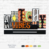 The Rustic Rhodes DIY Witchy Vibes Word Block features playful fonts and patterns—spiders, candy corn, checks—on a Halloween sign displayed on a white brick wall, with paint color options shown below for easy decorating.