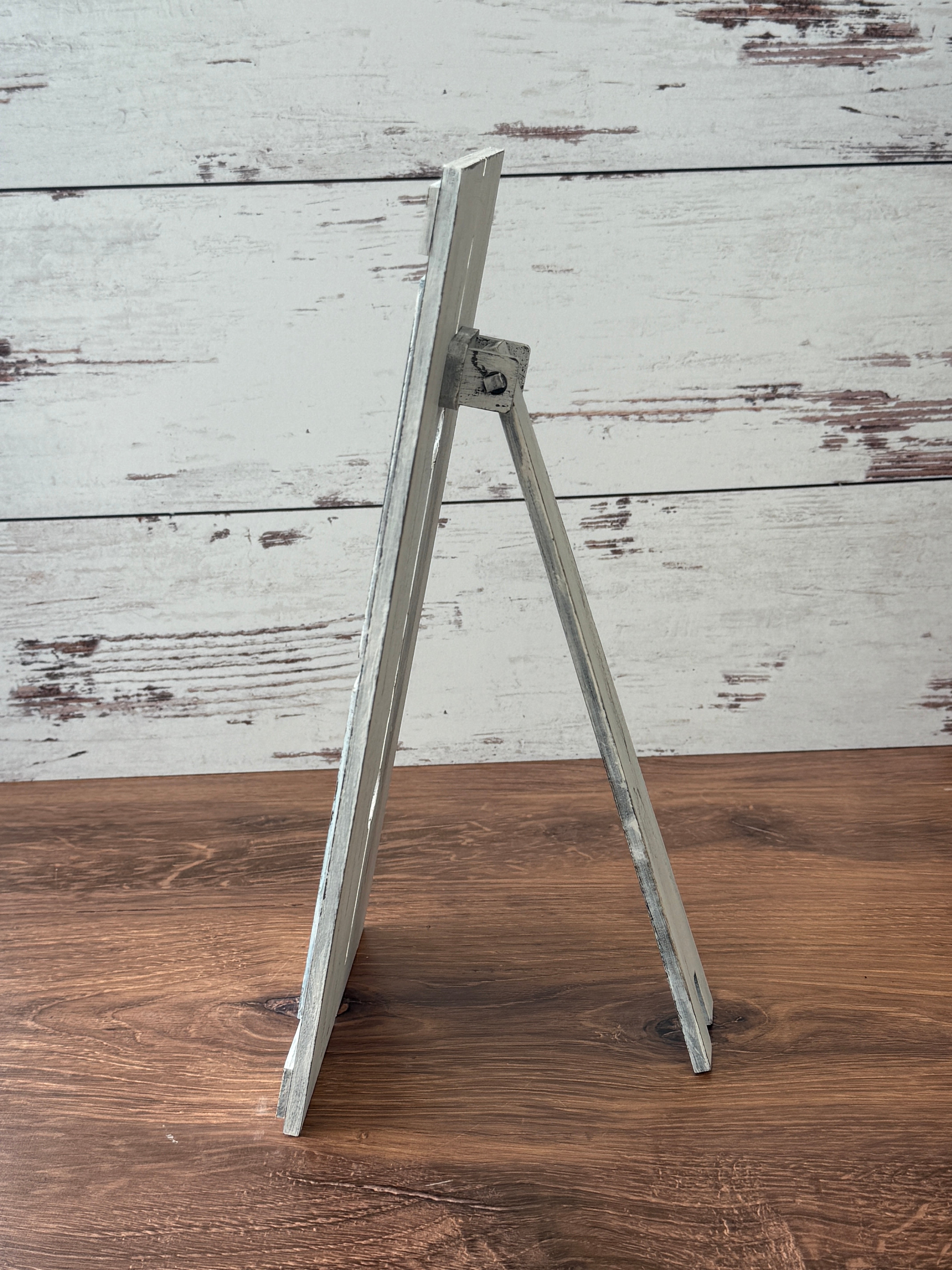 A small, rustic gray wood easel by The Rustic Rhode stands empty on a brown surface with a whitewashed background—ideal for displaying DIY Interchangeable Farmhouse Frame projects or custom decor. Side view shown.