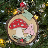 The Rustic Rhode's DIY Woodland Creatures Ornament- Mushrooms kit features a wooden ornament with red and pink mushrooms, greenery, a small red flower, plaid ribbon, and a "MERRY" tag—perfect for hanging on your lit Christmas tree.