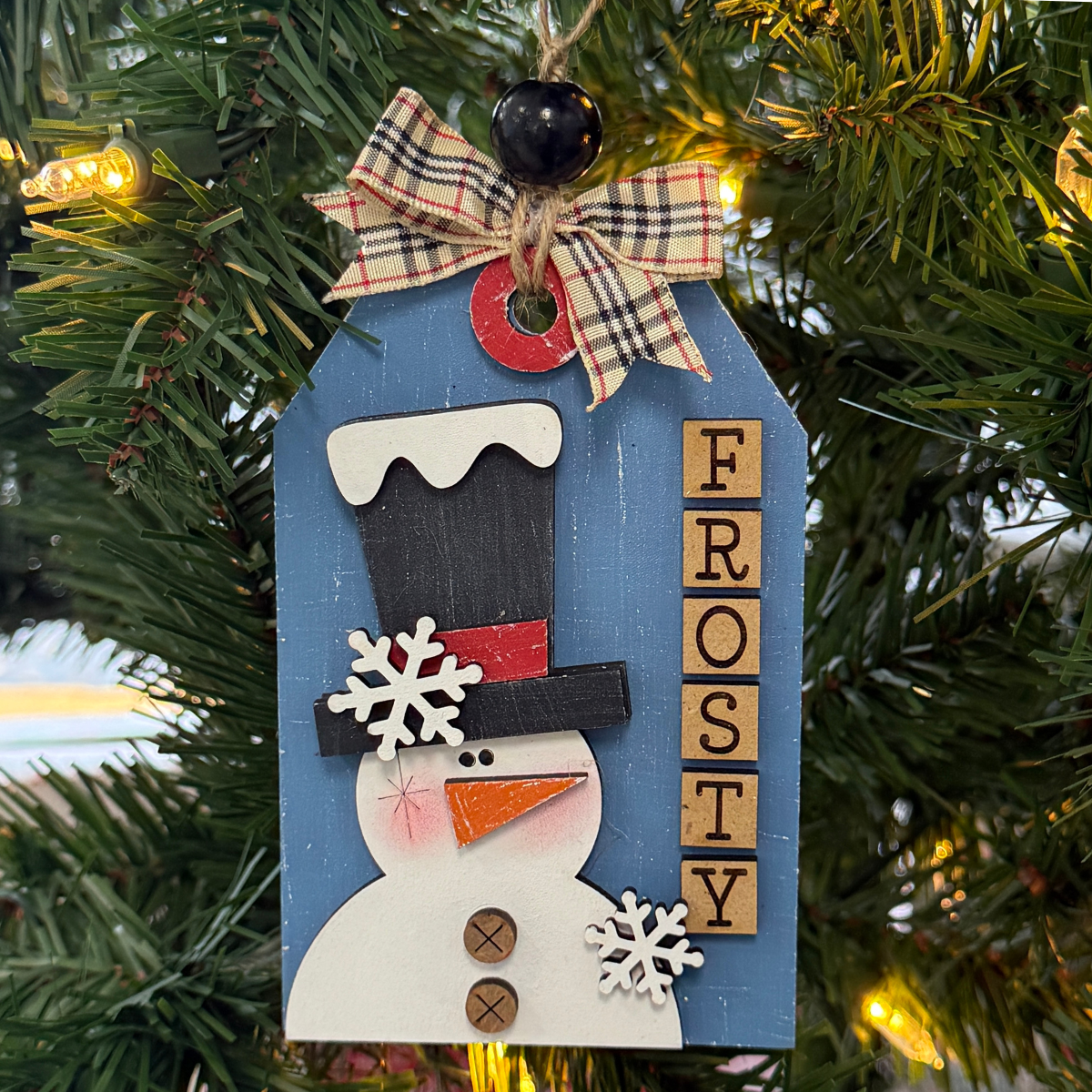 The Rustic Rhode’s DIY Prim Frosty the Snowman kit includes unassembled tag-shaped pieces with a snowman, black hat, plaid bow, snowflakes, and FROSTY in wooden blocks—paint before hanging on green pine branches and lights.