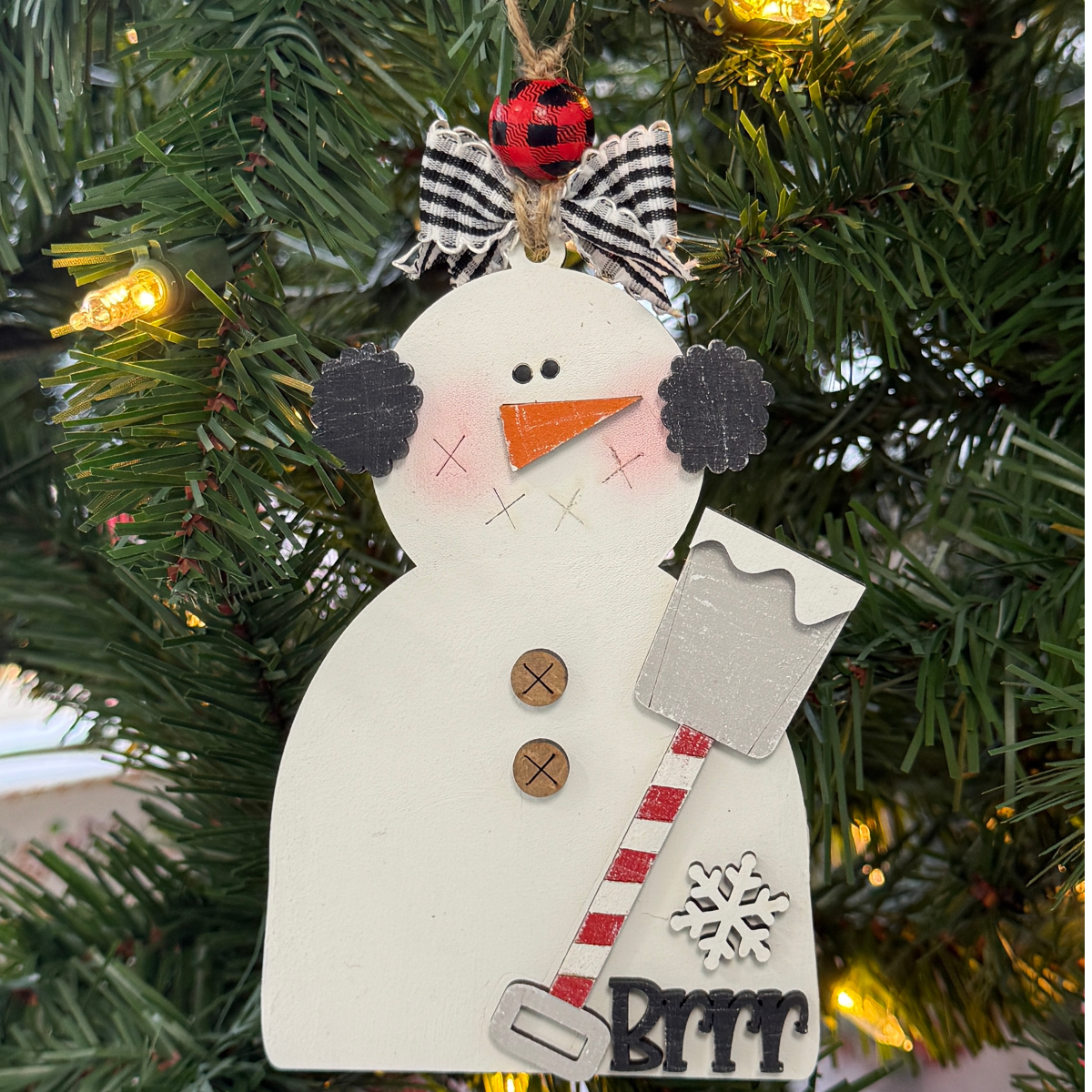 The Rustic Rhode's DIY Prim Snowman w/ Shovel kit includes unassembled pieces to create a snowman with rosy cheeks, button accents, a red-striped shovel, and a “Brrr” sign—perfect for glowing among warm Christmas lights.