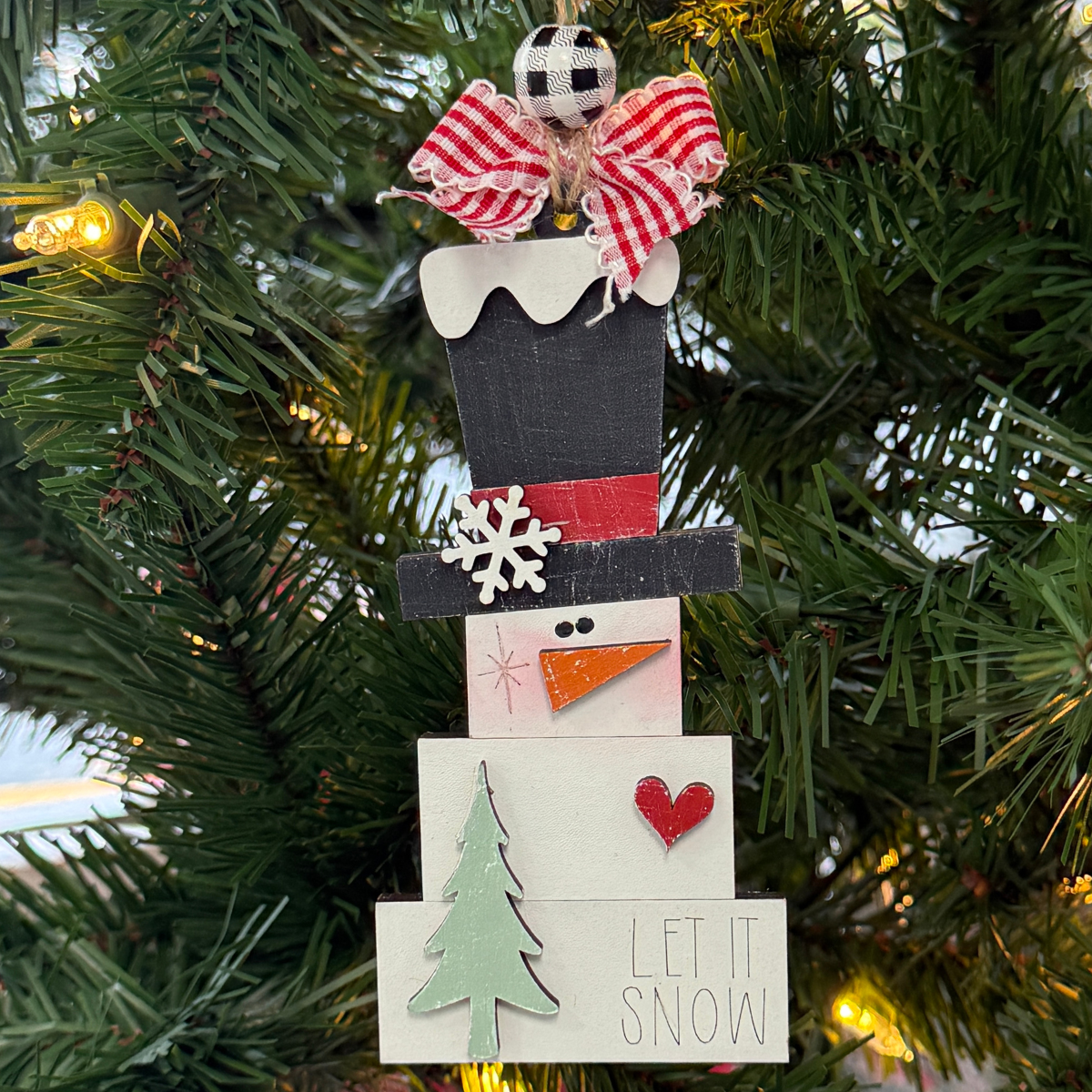 The Rustic Rhode DIY Prim Block Snowman ornament, ideal for ornament kits, features a top hat, red scarf, checkered bow, snowflake, red heart, green tree, and “Let it Snow” at the base. It hangs on a Christmas tree with glowing lights behind.