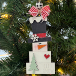 The Rustic Rhode DIY Prim Block Snowman ornament, ideal for ornament kits, features a top hat, red scarf, checkered bow, snowflake, red heart, green tree, and “Let it Snow” at the base. It hangs on a Christmas tree with glowing lights behind.