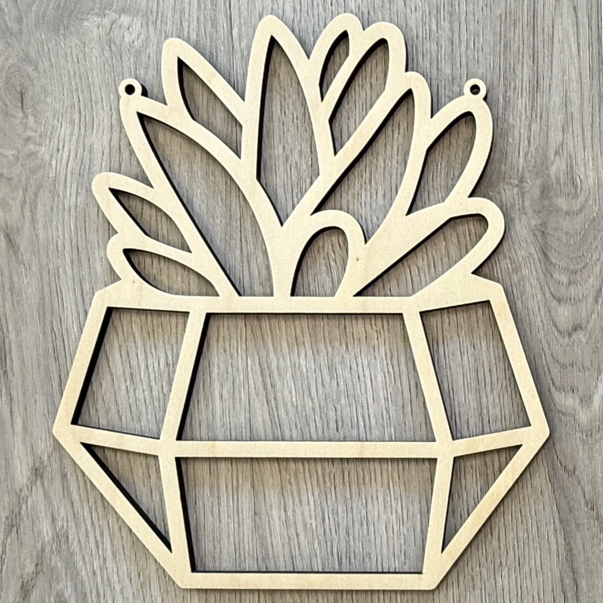The Rustic Rhode’s M. Mello Suncatcher Workshop- 10/7 features a geometric plant pot wooden cutout with stylized leaves and two top holes for hanging—perfect for DIY suncatchers or creative workshop projects.