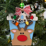 The Rustic Rhode’s DIY Christmas Latte-Rudolph is a reindeer ornament kit, featuring a red nose, colorful lights in its antlers, a red and white bow, and green checkered bead—perfect for painting or holiday crafting.