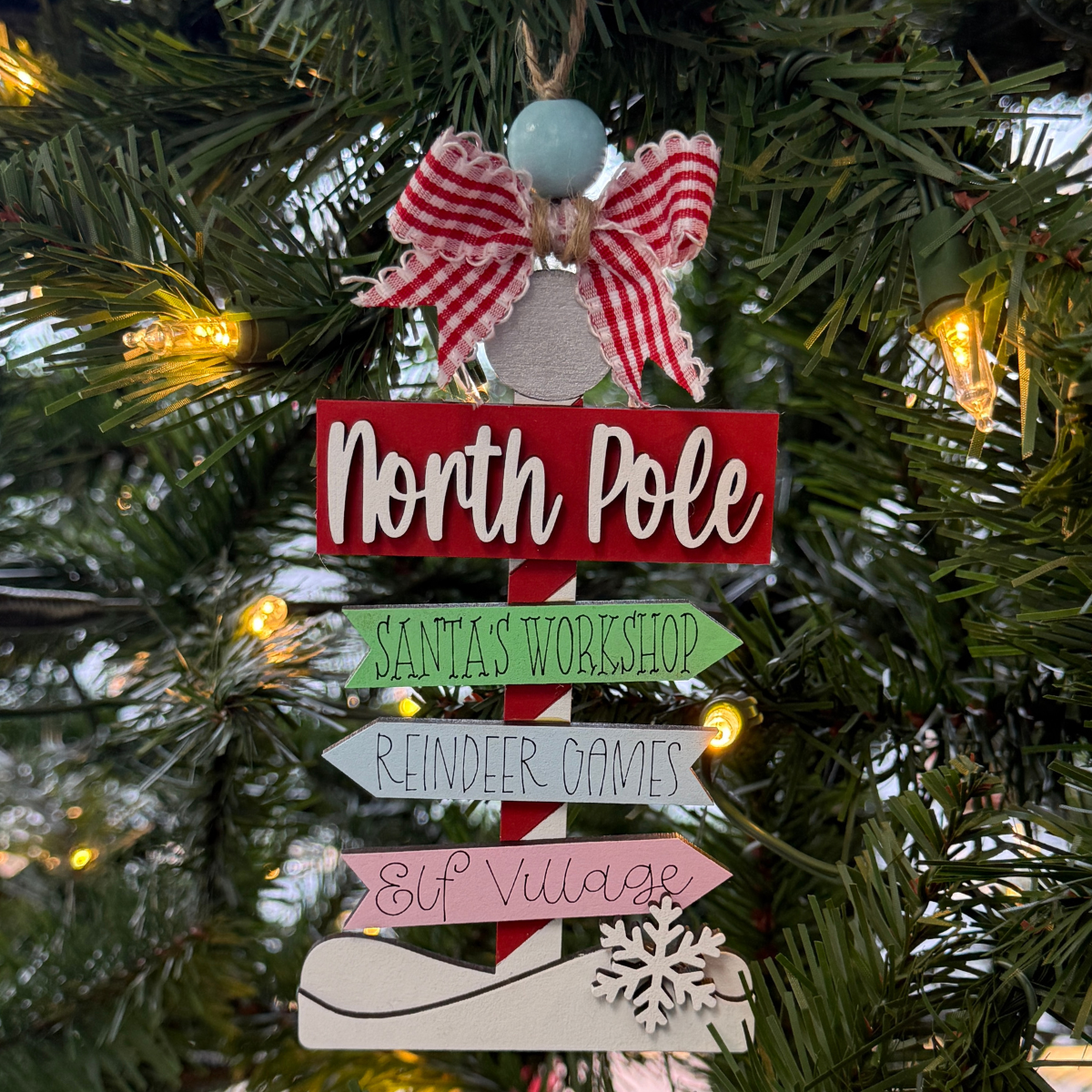 The Rustic Rhode's DIY North Pole Directional Ornament kit includes unassembled pieces to create a festive signpost with arrows to Santa's Workshop, Reindeer Games, and Elf Village, topped with a red-and-white bow and holiday accents.