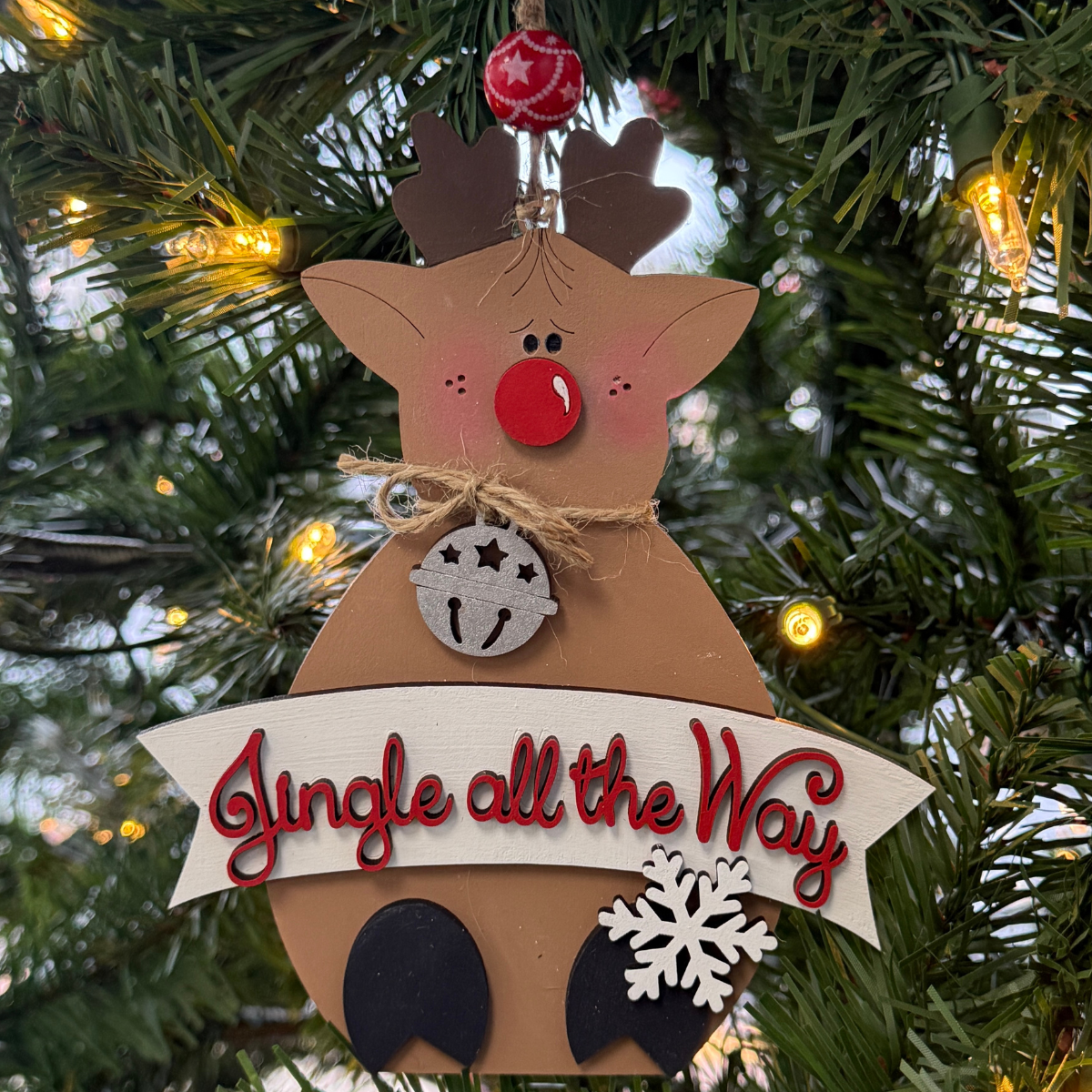 The Rustic Rhode’s DIY Rudolph Ornament features unassembled pieces to create a cute reindeer with a red nose, jingle bell, and snowflake. “Jingle all the Way” appears as it hangs on the Christmas tree with warm lights in the background.
