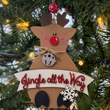 The Rustic Rhode’s DIY Rudolph Ornament features unassembled pieces to create a cute reindeer with a red nose, jingle bell, and snowflake. “Jingle all the Way” appears as it hangs on the Christmas tree with warm lights in the background.