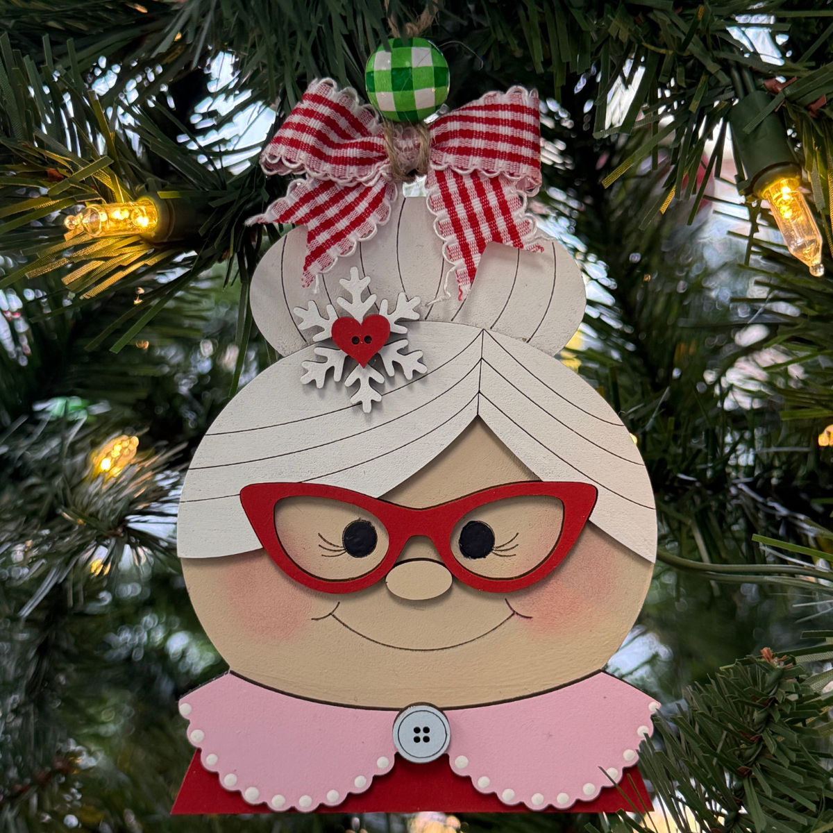 The Rustic Rhode DIY Mrs. Claus Ornament features red glasses, a snowflake hair accessory, and a red gingham bow, hanging on a Christmas tree among green branches and glowing yellow lights.