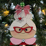 The Rustic Rhode DIY Mrs. Claus Ornament features red glasses, a snowflake hair accessory, and a red gingham bow, hanging on a Christmas tree among green branches and glowing yellow lights.