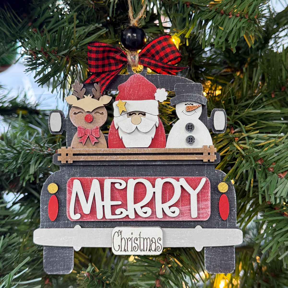 The Rustic Rhode DIY Truck Ornament- Santa's Friends kit features a black truck with "Merry Christmas," carrying a reindeer, Santa, and snowman. Includes unassembled pieces, red plaid bow topper, and green tree accents—just add paint!.