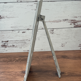 A small white wooden easel stands on a wood surface against a whitewashed plank background, displaying its side view—perfect for showcasing The Rustic Rhodes DIY Firefly Interchangeable 4.25 Insert.