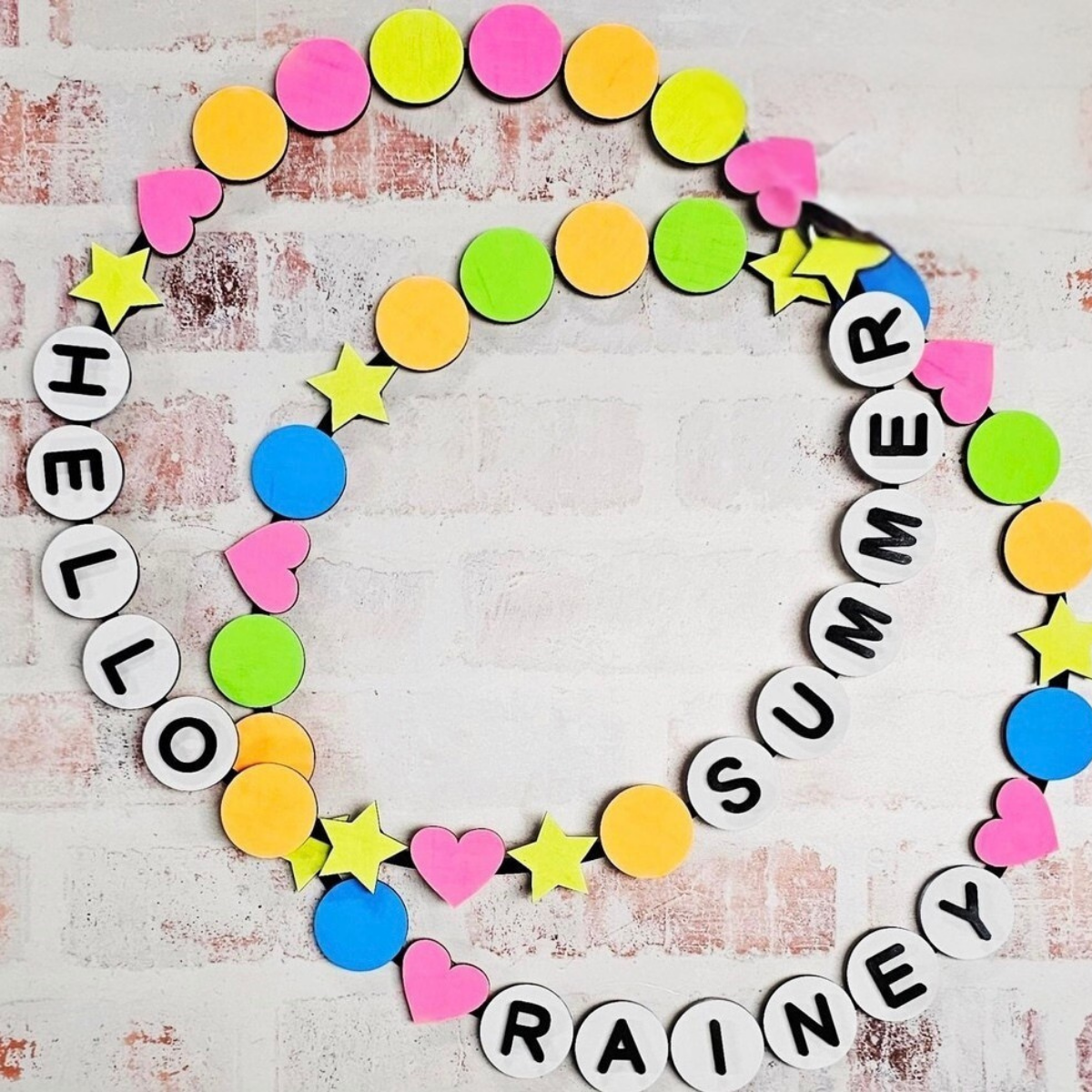 Colorful paper shapes (circles, hearts, stars) form a spiral on white brick, like a Friendship Bracelet Wreath. Lettered circles spell “Warwick Softball Fundraiser- 10/2/25” in pink, yellow, blue & green. Brand: The Rustic Rhode.