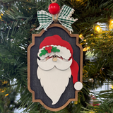 The Rustic Rhode DIY North Pole Santa Ornament kit includes unassembled wooden pieces of Santa with a red hat, white beard, and glasses. Finished with a green plaid bow and red bead, it hangs beautifully among pine needles and lights.