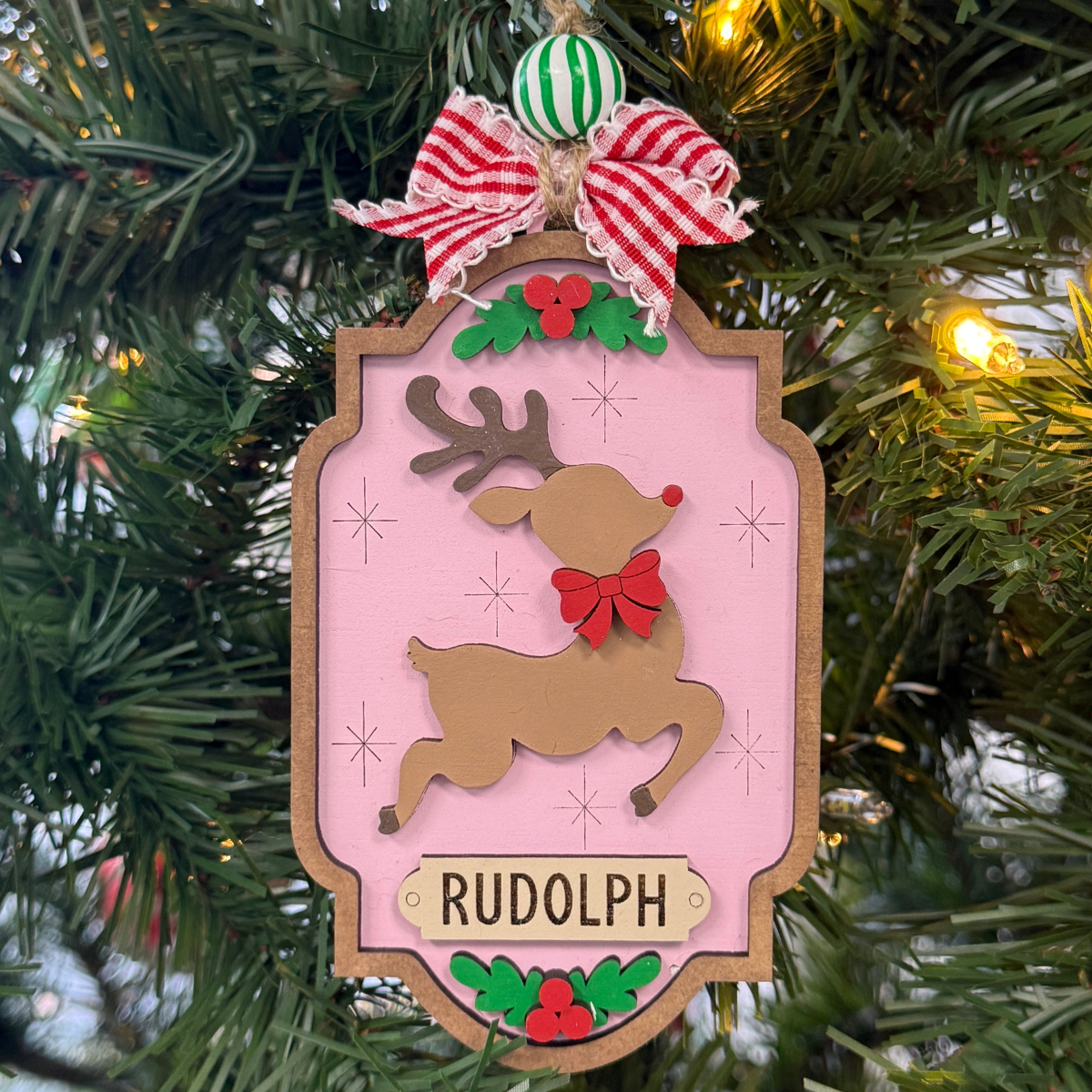 The Rustic Rhode DIY North Pole Rudolph Ornament kit features unassembled pieces, a striped ribbon, green and white bead, festive greenery, and red berries on a pink background—ready to paint and assemble with a reindeer and red bow design.