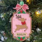 The Rustic Rhode DIY North Pole Rudolph Ornament kit features unassembled pieces, a striped ribbon, green and white bead, festive greenery, and red berries on a pink background—ready to paint and assemble with a reindeer and red bow design.
