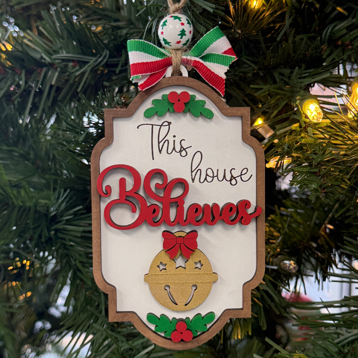 The Rustic Rhode's DIY North Pole This House Believes Ornament kit features a red bow, gold jingle bell, and red and green accents, perfect for hanging on your Christmas tree.