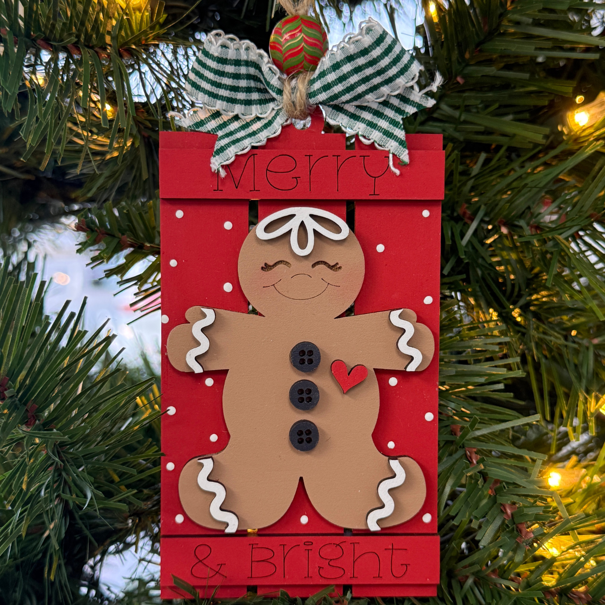 The Rustic Rhode's DIY Gingerbread Merry & Bright Pallet Ornament hangs on a Christmas tree, featuring a smiling gingerbread with black buttons, red heart, green plaid bow, and "Merry & Bright" as twinkling lights glow in the background.