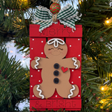 The Rustic Rhode's DIY Gingerbread Merry & Bright Pallet Ornament hangs on a Christmas tree, featuring a smiling gingerbread with black buttons, red heart, green plaid bow, and "Merry & Bright" as twinkling lights glow in the background.