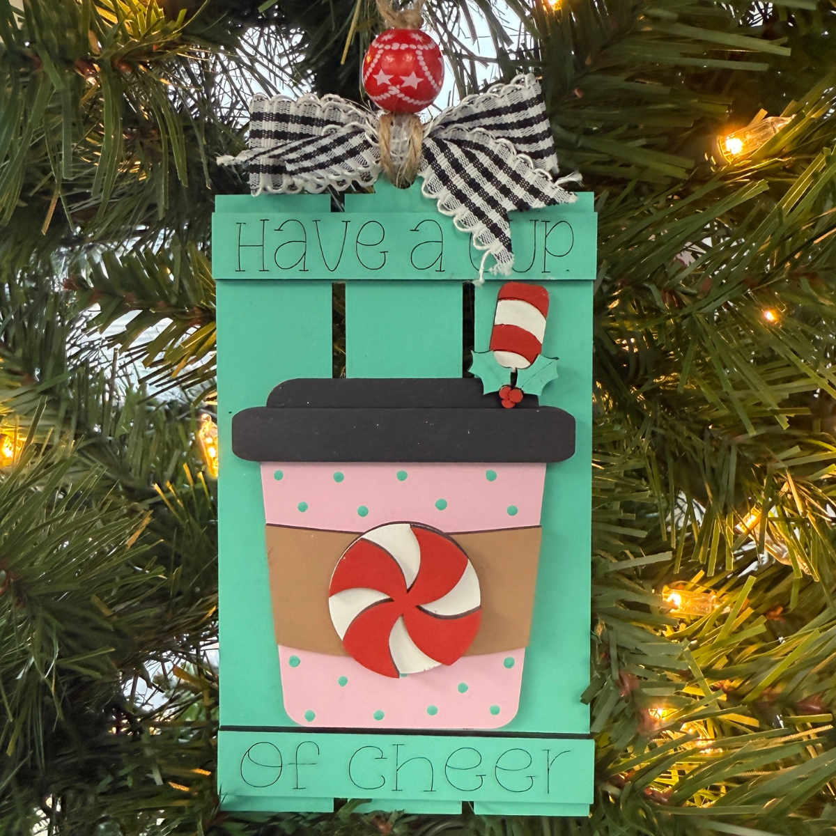 The Rustic Rhode’s DIY Hot Cocoa Pallet Ornament is a to-go cup-shaped Christmas tree kit with a mint candy, ribbon, and “Have a cup of cheer” on teal—perfect for painting and assembling yourself.