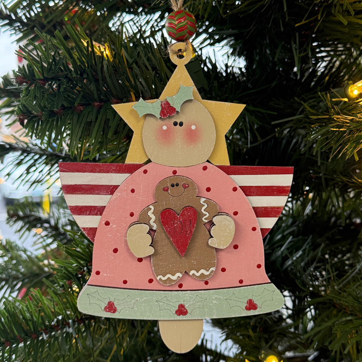 The Rustic Rhode's DIY Christmas Angel Ornament- Joy, painted pink with red stripes and holding a heart-decorated gingerbread cookie, hangs among pine needles and twinkling lights on the tree.