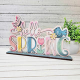 A colorful "hello SPRING" sign with flowers, a butterfly, and a blue bird from The Rustic Rhode by P. Fallon Group sits on a wooden table with a green potted plant in the background, perfect for your seasonal project menu.