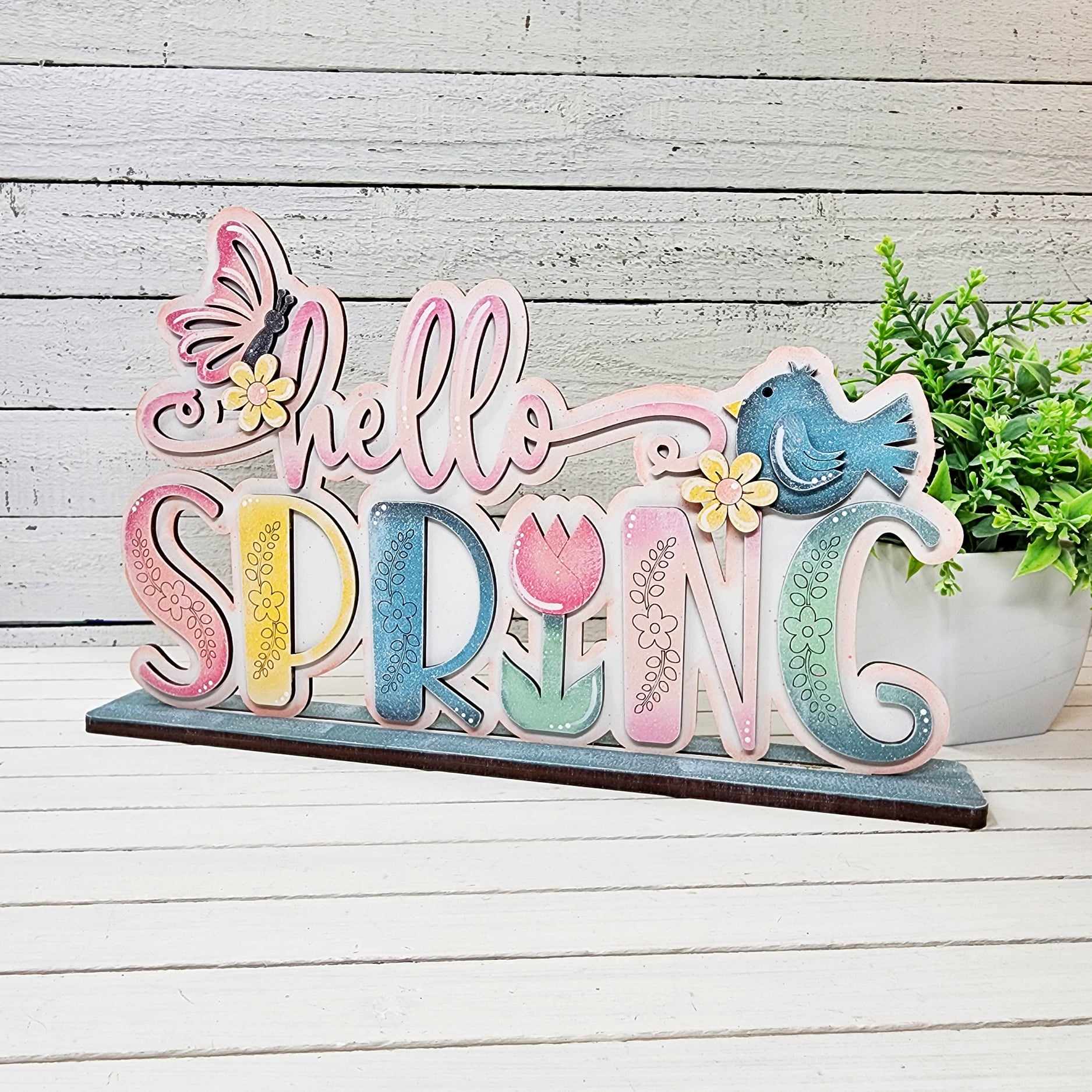 A colorful "hello SPRING" sign with flowers, a butterfly, and a blue bird from The Rustic Rhode by P. Fallon Group sits on a wooden table with a green potted plant in the background, perfect for your seasonal project menu.