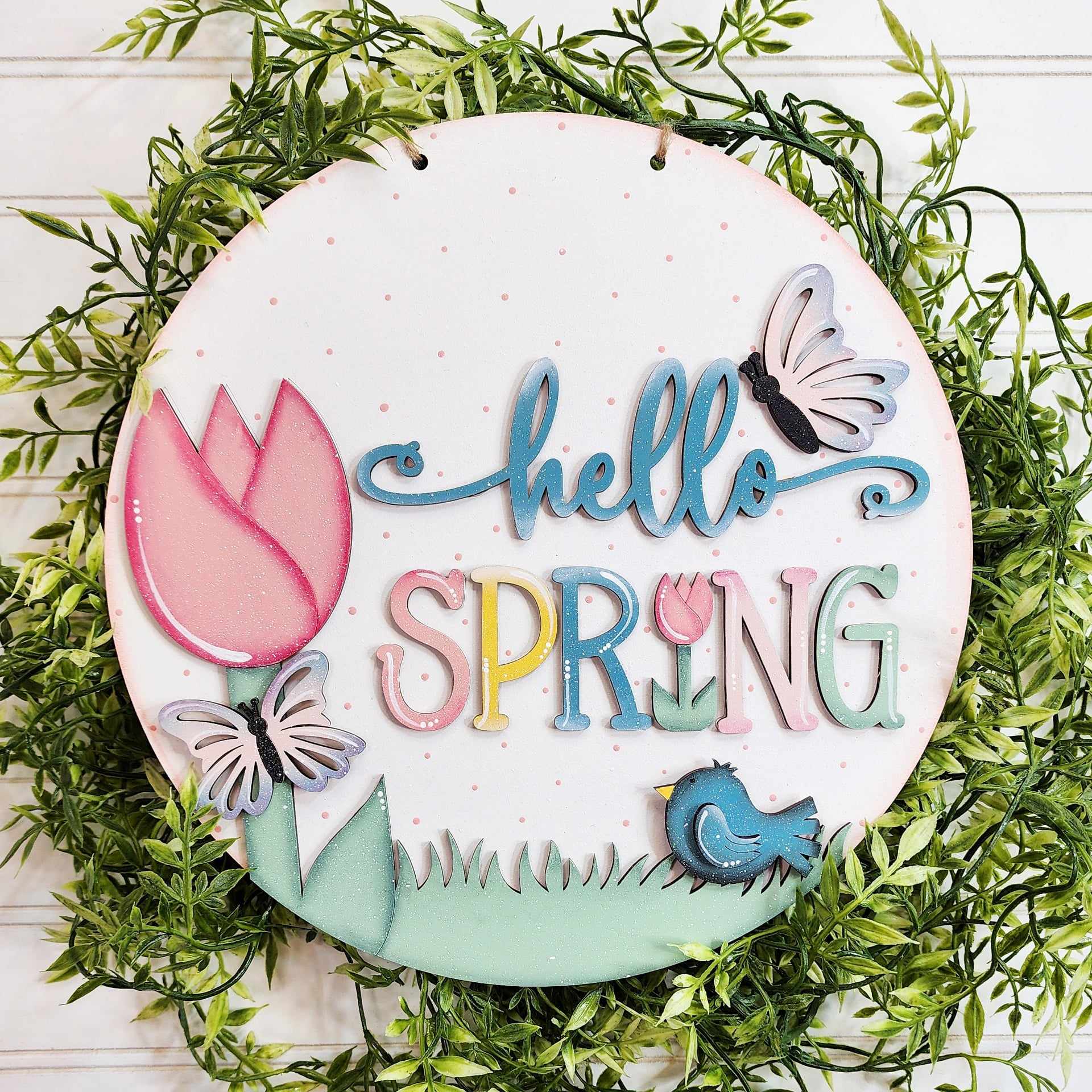 Add charm to your project menu with this round sign by The Rustic Rhode, featuring "hello SPRING" in colorful letters, a pink tulip, butterflies, a blue bird, grass, and green leaves on a white wood background from P. Fallon Group.