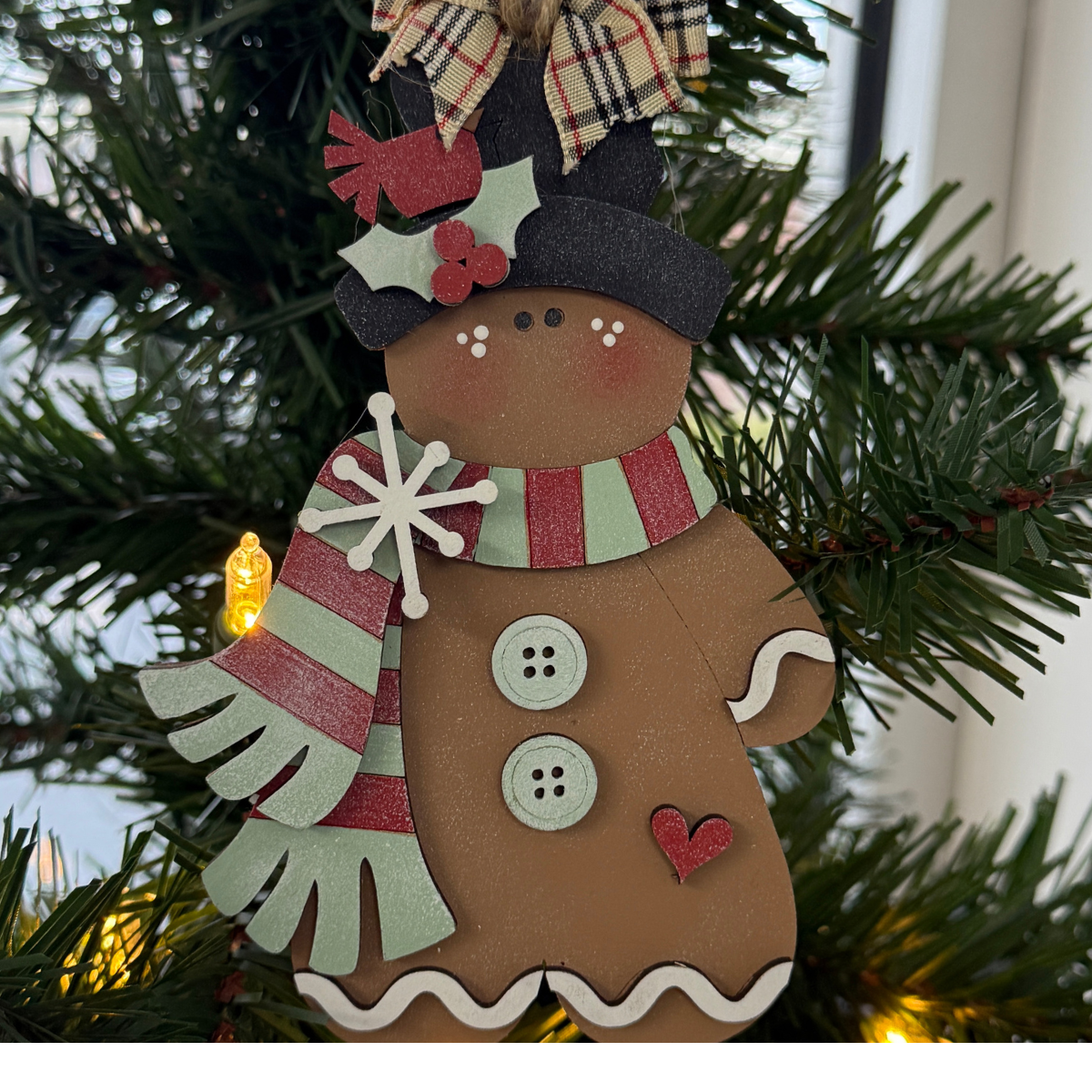 The Rustic Rhode DIY Victorian Gingerbread Scarf Ornament features a gingerbread person with a striped scarf, buttons, and heart detail, unassembled and shown hanging on a lit Christmas tree with greenery.