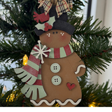 The Rustic Rhode DIY Victorian Gingerbread Scarf Ornament features a gingerbread person with a striped scarf, buttons, and heart detail, unassembled and shown hanging on a lit Christmas tree with greenery.