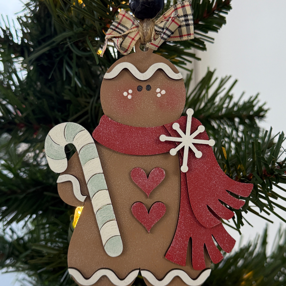 The Rustic Rhode's DIY Victorian Gingerbread Candy Cane Ornament features unassembled pieces for a gingerbread person with a red scarf, two red hearts, and a candy cane, hanging on a festive Christmas tree with green branches and a bow.